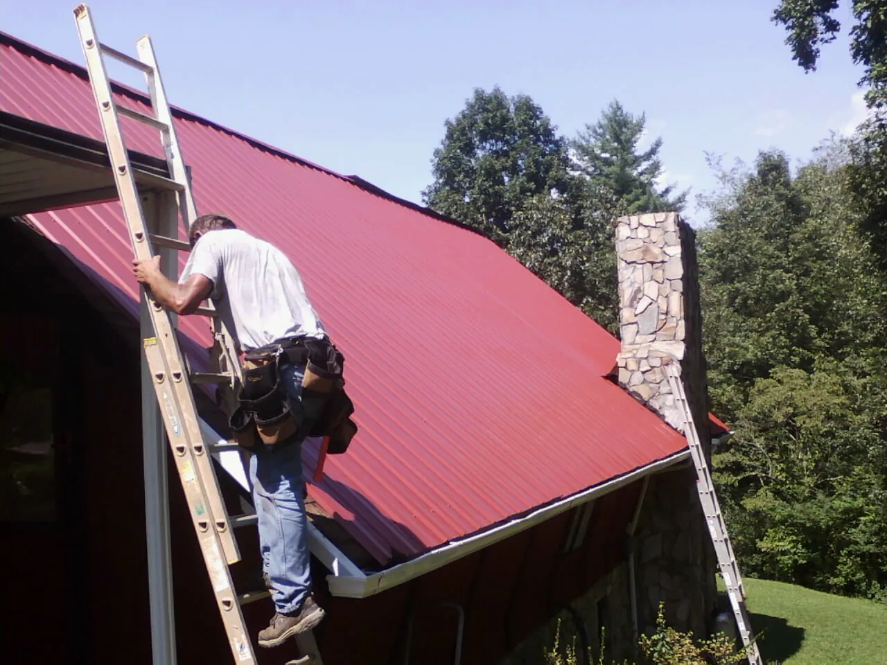 Roofer on a ladder beside a red metal roof and stone chimney — Soffit & Fascia Repair by Roofing Brothers in North Sandwich